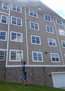 Man using a water-fed pole to clean windows on a multi-story building. The building has brown siding and stone accents. Cleaning windows helps to seize the perfect window of opportunity for a clear view.
