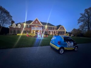Large brick house illuminated with festive lighting, showcasing holiday decorations on the lawn. An "Elite Power Washing" branded Smart Car is parked nearby, suggesting services for maintaining homes and holiday lighting.